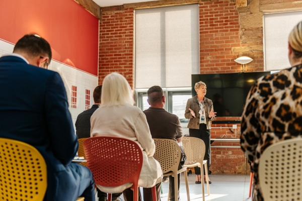 A person speaking at the front of a group of people. There are multiple rows of people sat watching the person speaking at the front of the room.