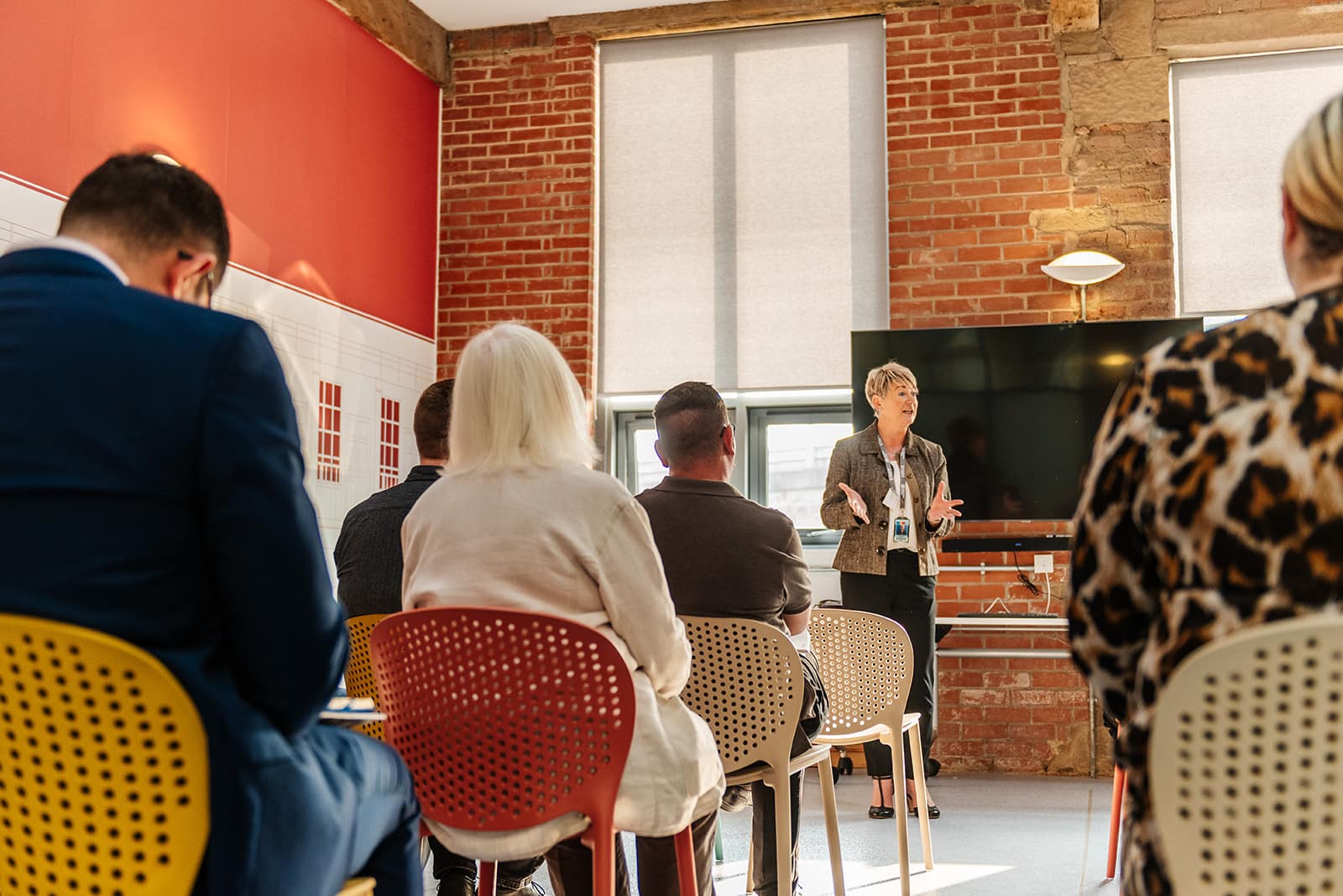 A person speaking at the front of a group of people. There are multiple rows of people sat watching the person speaking at the front of the room.