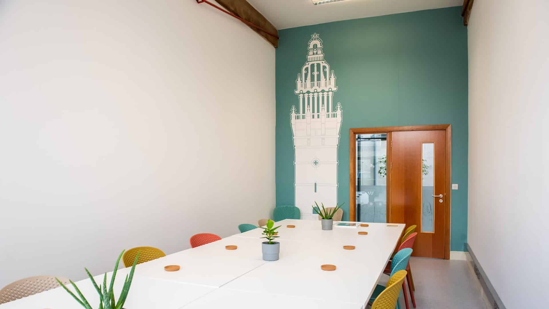 Long desk in a meeting room, with plants on the top. There is a bright blue wall in the background with a white mural on it.