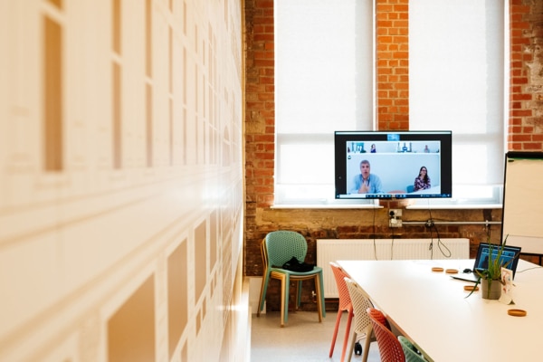 A meeting room with a tv screen, whiteboard and long white table. There is a virtual meeting happening on the screen.